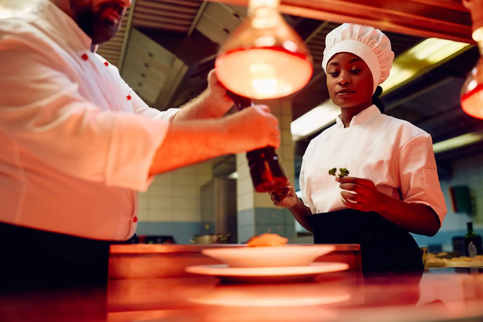 A chef plating a dish under heat lamps in a restaurant kitchen